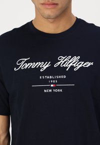 Homme portant un t-shirt bleu marine avec un texte blanc "Tommy Hilfiger Established 1985 New York" et un petit logo rouge, blanc et bleu.