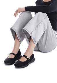 Zapatos Mary Jane de ante negro con punta redonda, correa ajustable con hebilla y suela de goma, combinados con pantalones de mezclilla gris de pierna ancha.