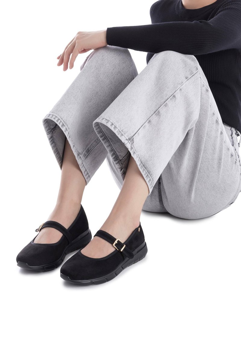 Zapatos Mary Jane de ante negro con punta redonda, correa ajustable con hebilla y suela de goma, combinados con pantalones de mezclilla gris de pierna ancha.