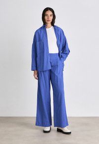Woman standing in blue pinstripe oversized shirt and wide-leg pants with white top and white platform shoes on gray floor.