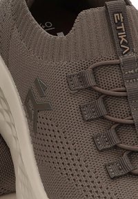 Acercamiento de una zapatilla deportiva de punto marrón con suela blanca, cordones elásticos y la marca "ETIKA" en el costado y en la lengüeta.