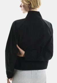 Musta lühike jope kõrge kraega, millel on sile tekstuur ja elastsed mansetid. Omab minimalismi ja raglanvarrukaid.