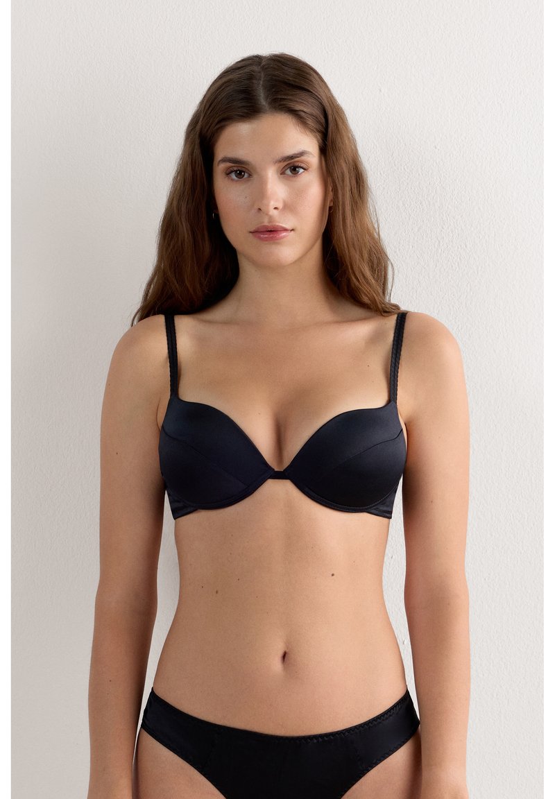 Femme aux longs cheveux bruns portant un soutien-gorge noir à armatures et une culotte noire assortie devant un mur blanc uni.