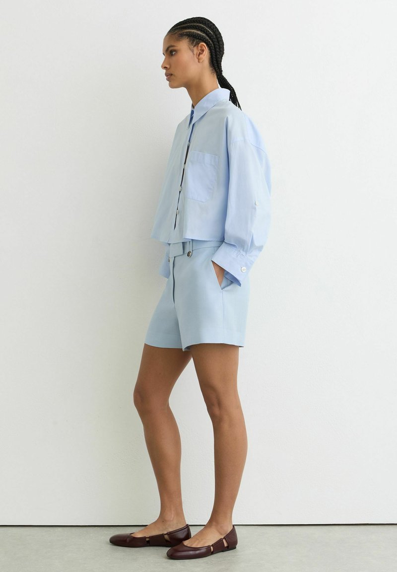 Žena s vrkočmi, oblečená v svetlomodrej košeli na gombíky a zodpovedajúcich šortkách, stojaca z profilu s rukami v vreckách a hnedými balerínami.