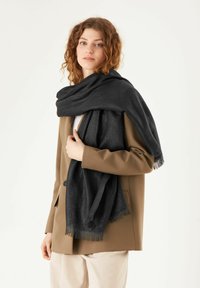 Zwarte sjaal gemaakt van een zachte, gestructureerde stof. Het heeft franjes aan de randen en is om de schouders van een persoon gewikkeld, ter aanvulling op een beige jas.