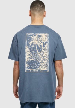 Camiseta de algodón azul con un gráfico color crema de palmeras y olas del océano en la parte posterior, con el texto "E KOMO MAI" en la parte inferior.