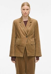 Blazer en velours côtelé marron avec deux poches avant, revers crantés et fermeture à un bouton ; coupe près du corps avec une surface texturée.
