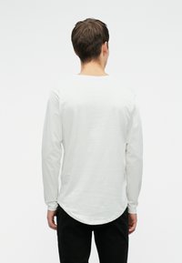 Mand med kort brunt hår iført en hvid langærmet skjorte og sorte bukser, stående med ryggen til kameraet foran en ensfarvet baggrund.