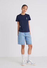 Mørkeblå bomull t-skjorte med små blomsterbroderier på venstre bryst, kombinert med lyseblå denimshorts og grå joggesko.