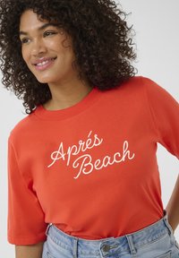Chemise rouge à manches courtes avec un texte en script blanc "Apres Beach". Tissu en coton doux, encolure ronde, assortie à un short en denim taille haute.
