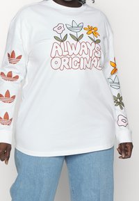 Vit långärmad skjorta med "Always Original" i fet stil, blommönster på ärmarna och Adidas-logotypdetaljer i rött.
