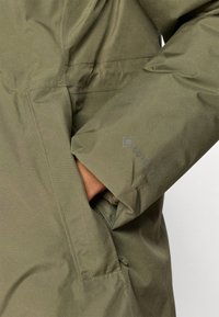 Olivově zelená voděodolná bunda s detailním záběrem na boční kapsu a texturovanou tkaninou, s viditelným logem "GORE-TEX" na rukávu.