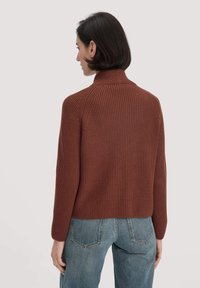 Brauner Strickpullover mit hohem Kragen und gerippter Textur, mit einem cropped Schnitt und langen Ärmeln, kombiniert mit hellblauen Jeans.
