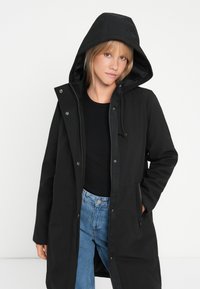 Black hooded raincoat, features snap button closure, adjustable drawstring hood, and side pockets. Worn over a black top and blue jeans.