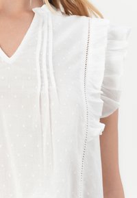 Weißes Blusenshirt mit V-Ausschnitt, das mit Längsplissees, gestickten Akzenten und Rüschensäumen verziert ist. Strukturiertes Gewebe mit kleinen Punktmustern.