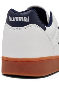 Fehér sneaker tengerészkék sarokkal, textúrált gumi talppal. Az anyag sima szintetikus, kontrasztos varrással. A márka logója látható.
