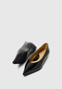 Chaussures noires en cuir à bout pointu avec un talon bas et des découpes. Texture lisse avec une finition brillante et une semelle intérieure douce et rembourrée.