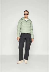Vrouw in een lichtgroene puffer jack, zwarte cargobroek, beige sneakers en zonnebril, staand tegen een effen lichte achtergrond.