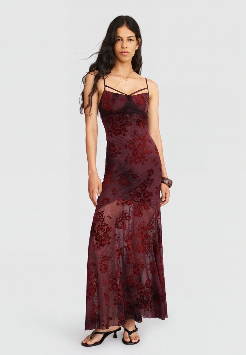 Woman with long dark hair wearing a floor-length burgundy dress with floral pattern and thin straps, standing with one hand relaxed.