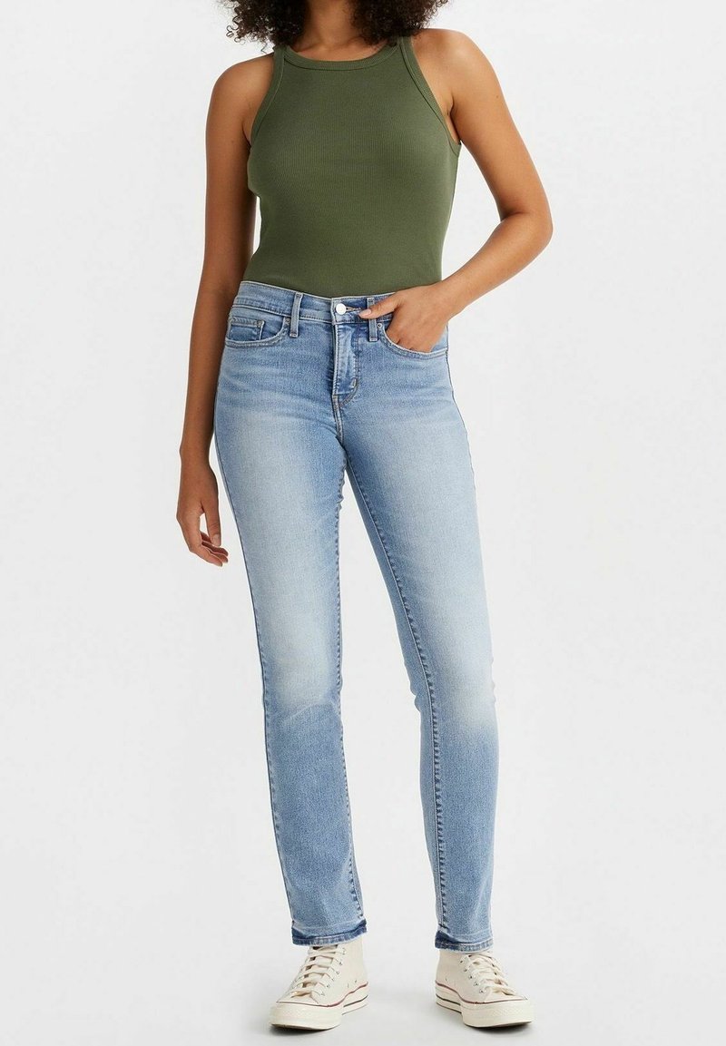 Femme portant un haut sans manches vert olive, un jean taille haute bleu clair et des baskets blanches, debout, une main dans la poche.