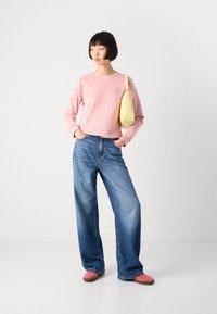 Vrouw staat met handen in de zakken, draagt een roze sweatshirt, wijde blauwe jeans, roze schoenen en een gele schoudertas op een witte achtergrond.