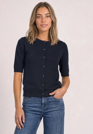 Femme portant un cardigan noir à manches courtes boutonné et un jean bleu, debout avec une main dans la poche, sur un fond clair.