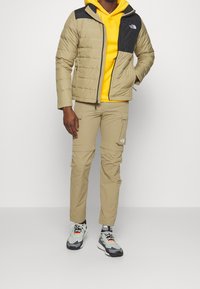 Veste beige matelassée avec panneau d'épaule noir, associée à un sweat à capuche jaune et un pantalon cargo beige. Chaussures grises et noires avec des accents orange.