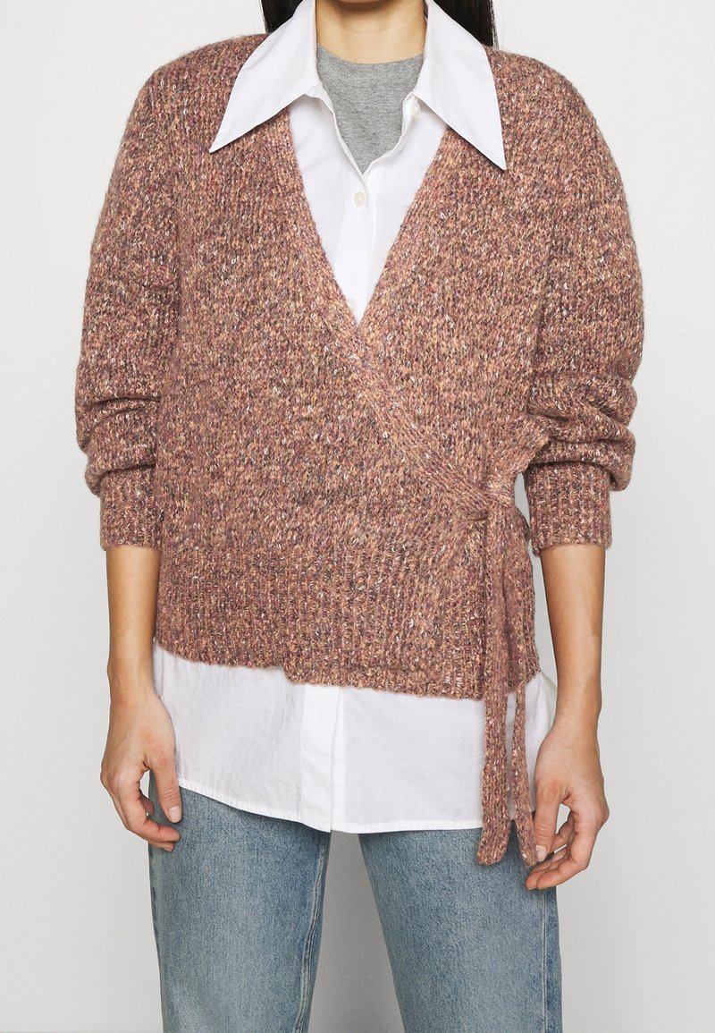 Personne portant un pull rose texturé en cache-cœur par-dessus une chemise blanche boutonnée et un sous-vêtement gris avec un jean bleu, bras détendus le long du corps.