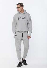 Jasnoszara bluza z kapturem z czarnym logo w formie napisu oraz dopasowane spodnie dresowe, z czarnymi sznurkami ściągającymi i kieszeniami zapinanymi na zamek, w zestawie z czarnymi sneakersami.