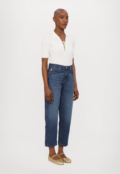 Top blanco de manga corta con costuras en relieve y cordones en el frente, combinado con unos vaqueros de denim de tiro alto en azul y unas chanclas doradas de tejido.