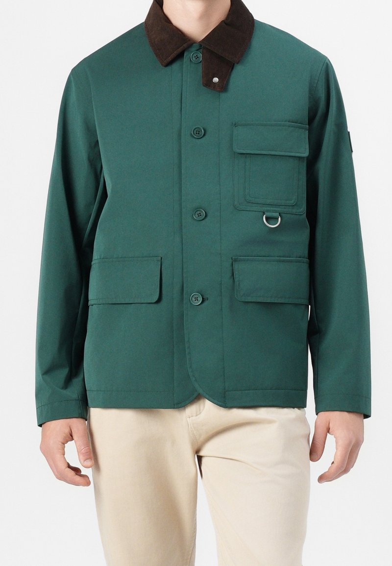 Homme portant une veste verte boutonnée avec un col marron foncé et plusieurs poches avant, associée à un pantalon beige.