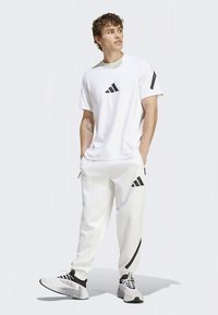 Balta sportinė apranga, kurioje yra trumpomis rankovėmis marškinėliai ir siaurėjančios kelnės, abu su juodais adidas logotipais ir akcentais. Vaizdą papildo tekstūruoti sportbačiai.