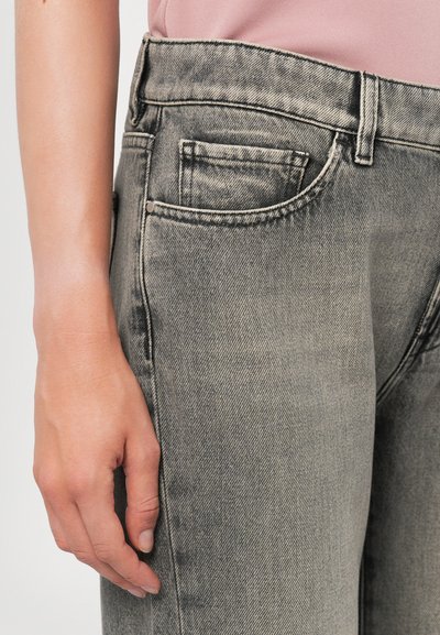 Jeans de mezclilla gris de alta cintura, con un pequeño bolsillo frontal, costuras visibles y una textura descolorida. Mano posicionada en el costado.