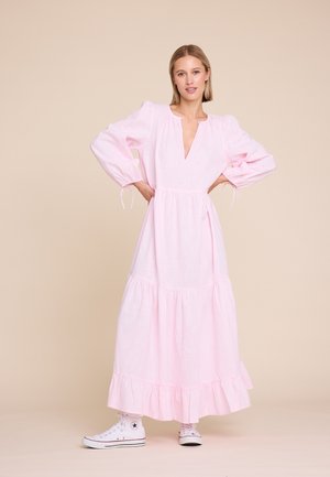 Kvinne iført en lang, lys rosa kjole med puffede ermer, kombinert med hvite høye sneakers, stående med hendene på hoftene.