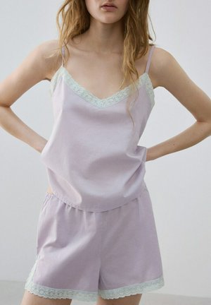 Kobieta w jasnofioletowym topie na ramiączkach z koronką i dopasowanych szortach, stojąca z rękami na biodrach na jednolitym tle.