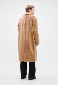 Brūns mākslīgās ādas mētelis ar plašu apkakli un garām piedurknēm, kas sasniedz puslīdz potītēm, uzvilkts virs melnajiem biksēm un zābakiem. Ar teksturētu un pūkainu izskatu.