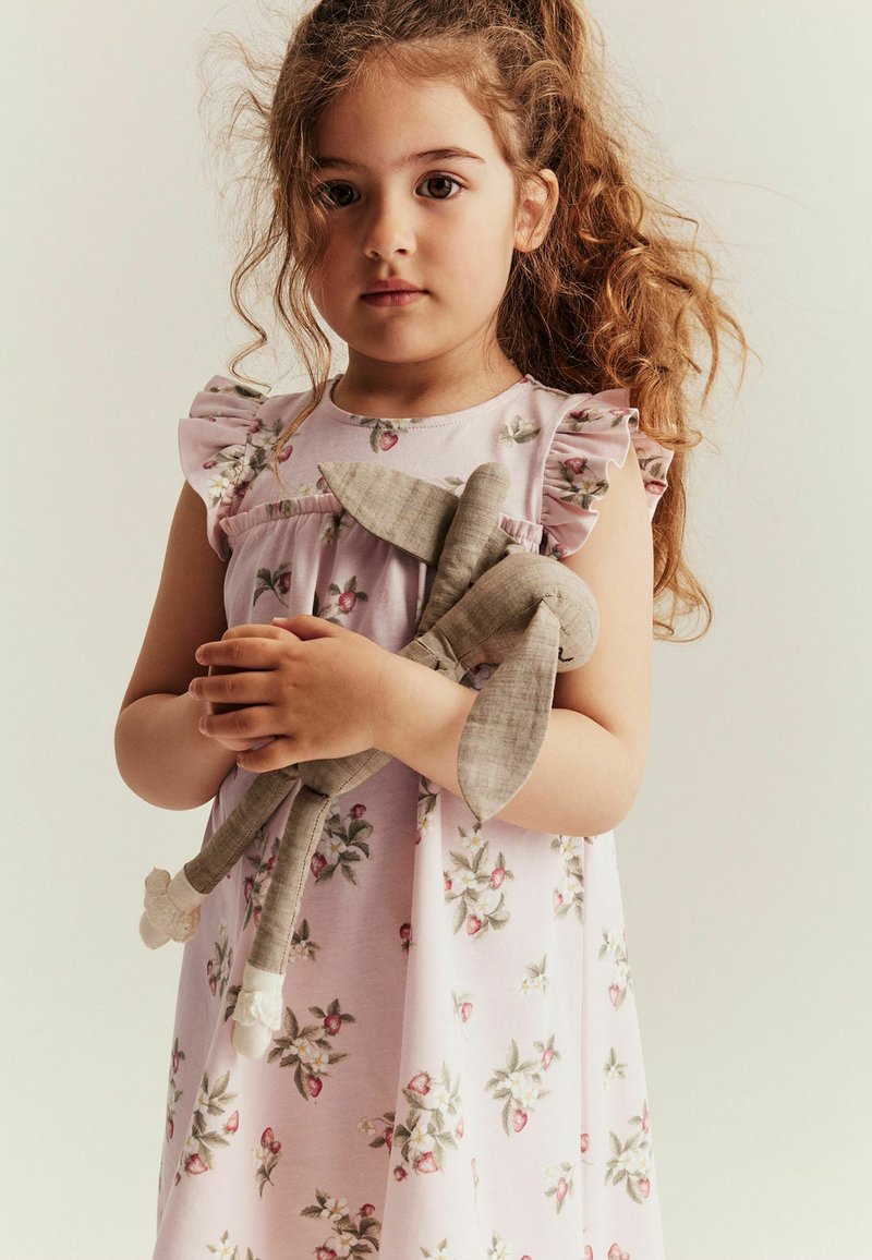 Hellrosa Kleid mit Blumenmuster, Rüschensäumen und einer weichen Textur; das Kind hält ein beiges LeinenkaninchenSpielzeug mit langen Ohren.