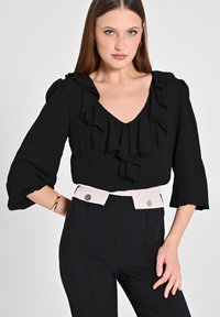 Blusa nera con scollatura arricciata e maniche a tre quarti, abbinata a pantaloni a vita alta con un cinturino di colore chiaro e bottoni decorativi.
