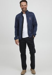 Dunkelblaue Zip-Jacke mit strukturierter Oberfläche, schwarzem Besatz und Logoschriftzug. Kombiniert mit Cargo-Hosen und beigen Stiefeln.