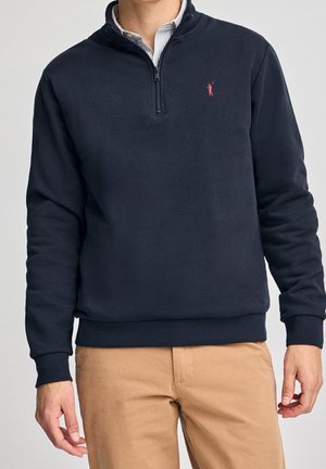 Muž v tmavě modré mikině na čtvrtý zip s malým červeným logem na hrudi a v béžových kalhotách, stojící s uvolněnými pažemi.