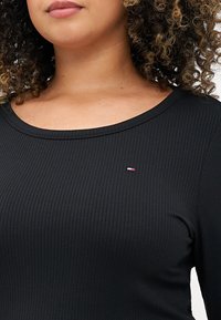 Schwarzes, geripptes Langarmoberteil mit Rundhalsausschnitt, mit einem kleinen Logo in Rot, Weiß und Blau auf der Brust. Weicher Stoff und taillierte Passform.