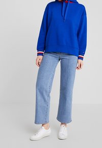 Modrá mikina s kapucí v uvolněném střihu, s proužkovanými manžetami v červené a bílé barvě. Kombinována s širokými světlými modrými džínami a bílými teniskami.