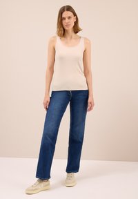 Donna in piedi che indossa un top smanicato beige, jeans dritti blu e sneakers beige su uno sfondo semplice.