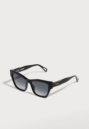Lunettes de soleil noires forme œil de chat avec des verres à dégradé foncé et de petits clous dorés le long des branches sur un fond clair.
