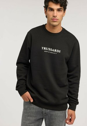 Joven con sudadera negra con texto ""TRUSSARDI GENTLE SOCIETY"", combinado con vaqueros grises, fondo neutro.