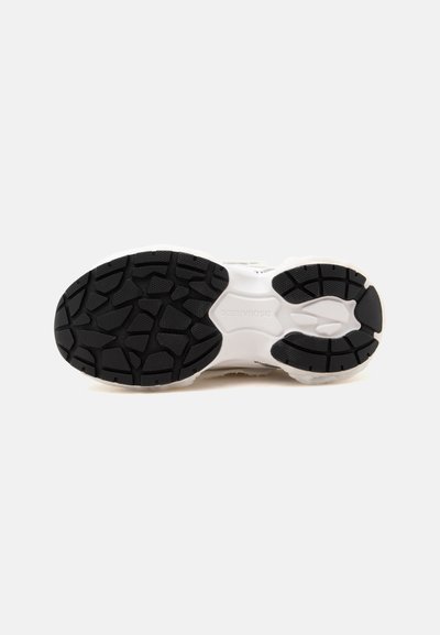Semelle d’une basket blanche avec des patins en caoutchouc noir pour l’adhérence et un motif de bande de roulement texturé, montrée sur un fond clair uni.