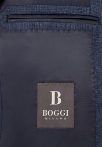 Jūras spēku audums ar tekstūru un gludu apdari; ar brūnu etiķeti, uz kuras ir logo "B" un teksts "BOGGI MILANO."