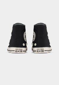 Μαύρα ψηλά αθλητικά παπούτσια Converse All Star, διακοσμημένα με λευκά μοτίβα μαργαρίτας, σε απλό φόντο, με θέα από πίσω.
