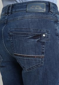 Nära bild av bakfickan på mörkblå denimjeans med en sydd dekorsömm och en läderetikett på midjan.