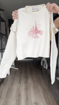 Sudadera recortada de color crema con mangas largas, que presenta un gráfico rojo de la Torre Eiffel y el texto 'París', con detalles de diseño en las mangas enrolladas.